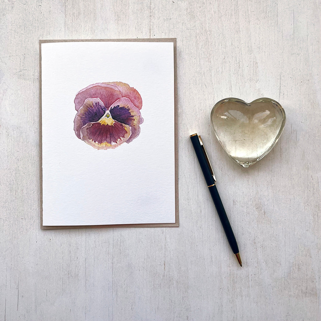 A note card featuring a delicate watercolour painting of a burgundy pansy with hints of pink and yellow. Artist Kathleen Maunder.