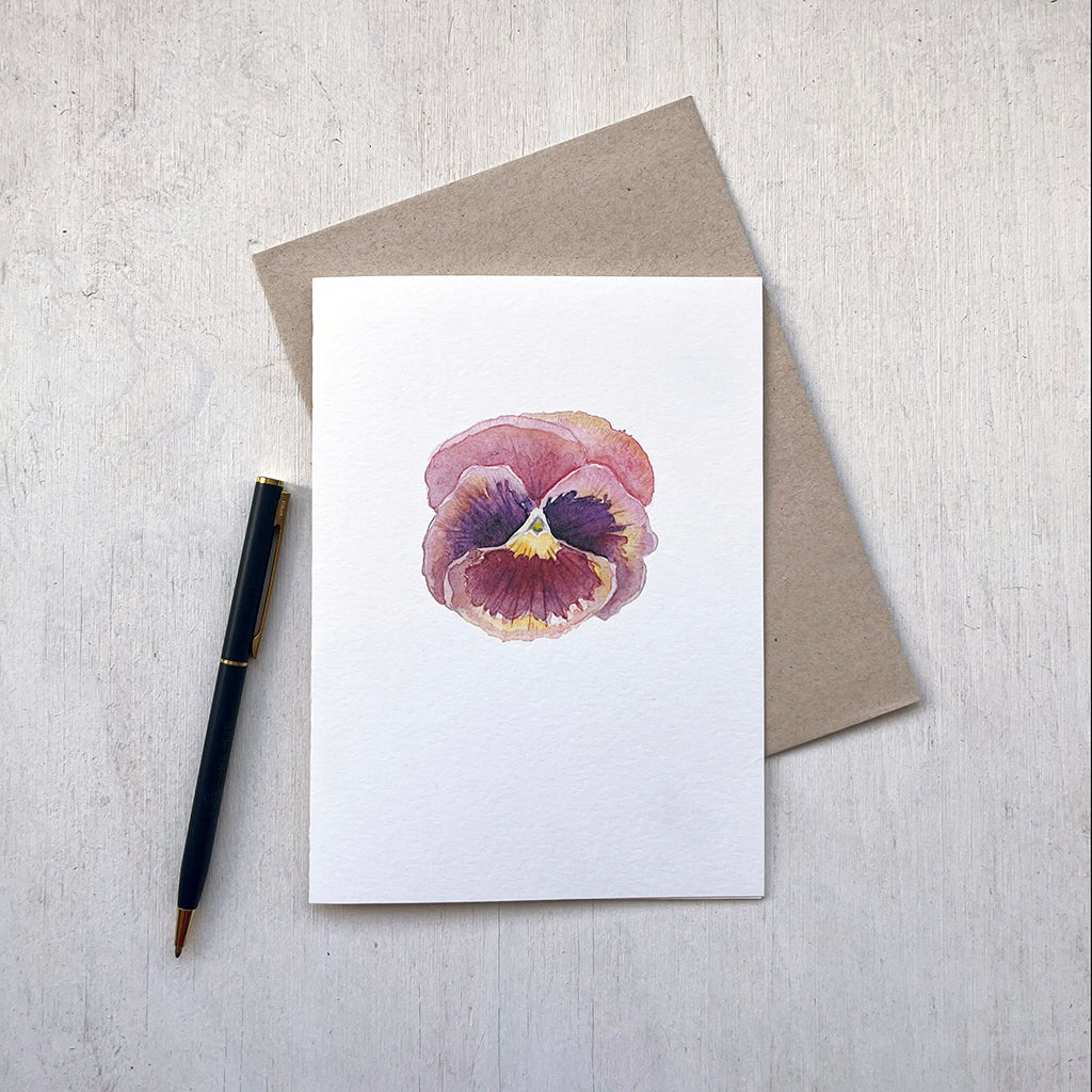 A note card featuring a watercolor painting of a single burgundy pansy with it accompanying recycled envelope. Artist Kathleen Maunder