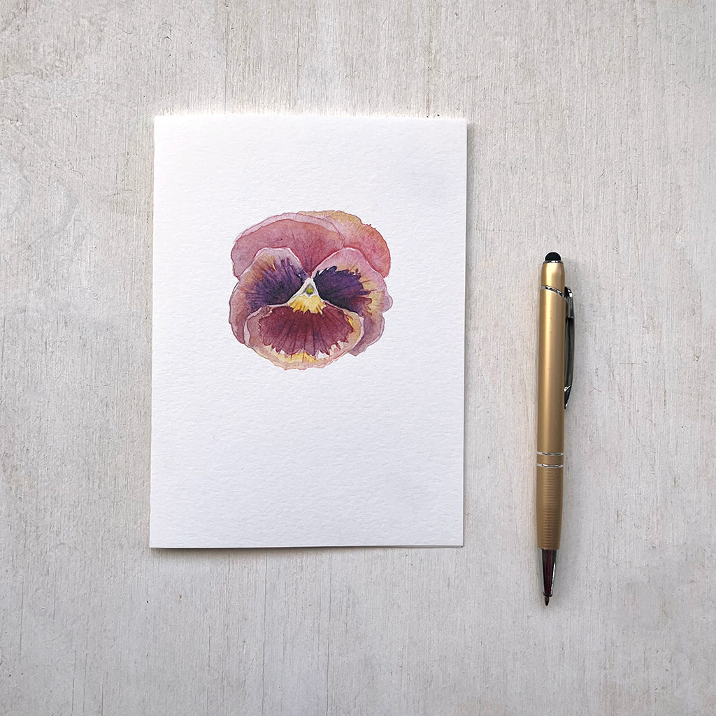 A note card featuring a beautiful watercolour painting of a burgundy pansy with tones of pink and yellow. 