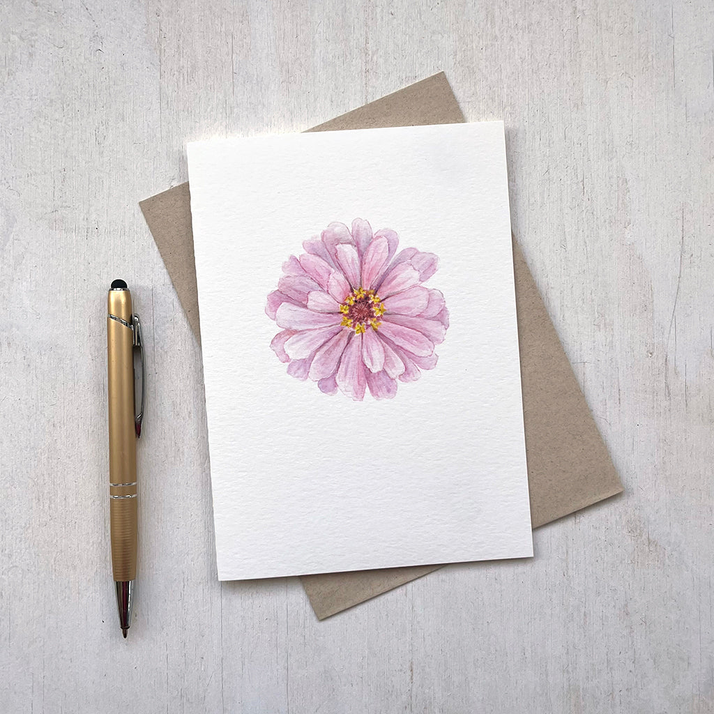 A blank note card features a lovely watercolour painting of a pink zinnia on its front. Its accompanying recycled envelope  sits behind it.