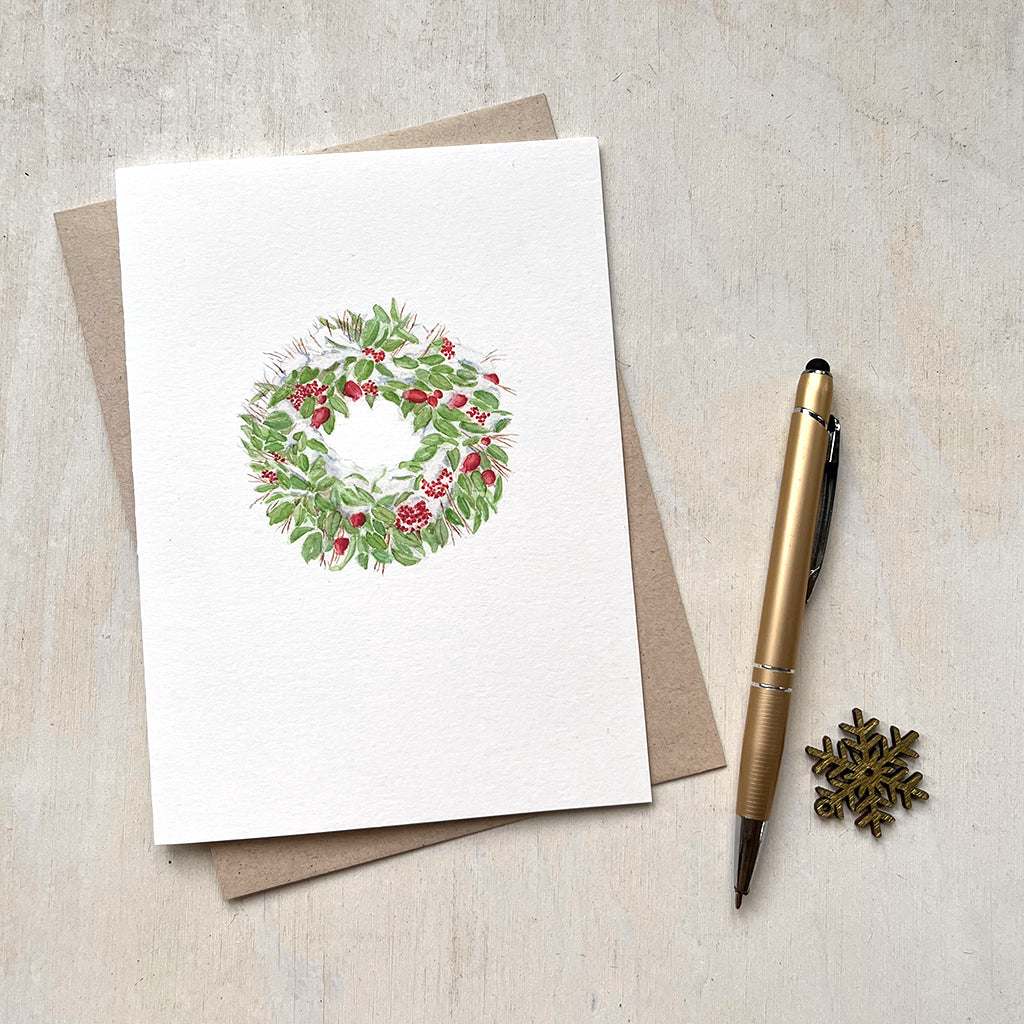 A holiday card with a delicate watercolour painting of a snow-covered Christmas wreath made of green leaves, red fruit and berries.