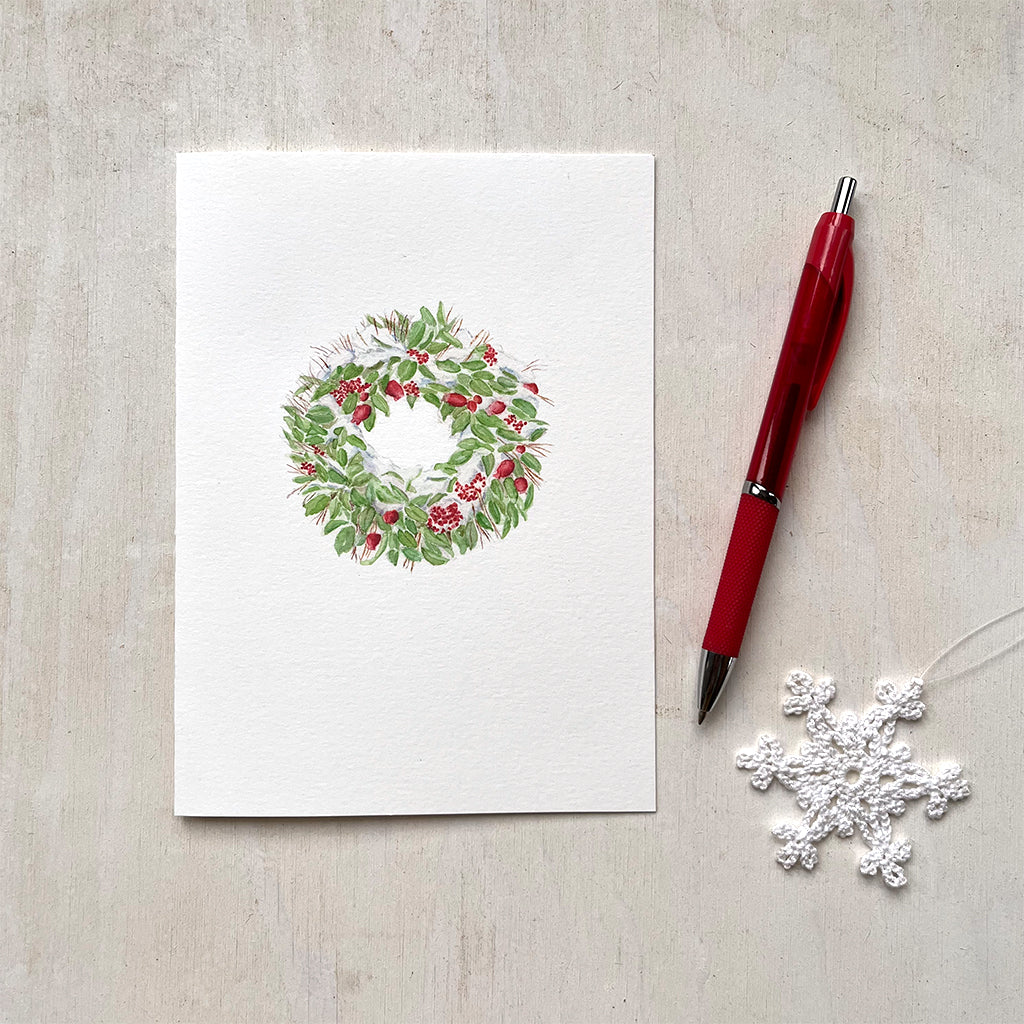 A holiday card with a delicate watercolour painting of a snow-covered Christmas wreath made of green leaves, red fruit and berries.