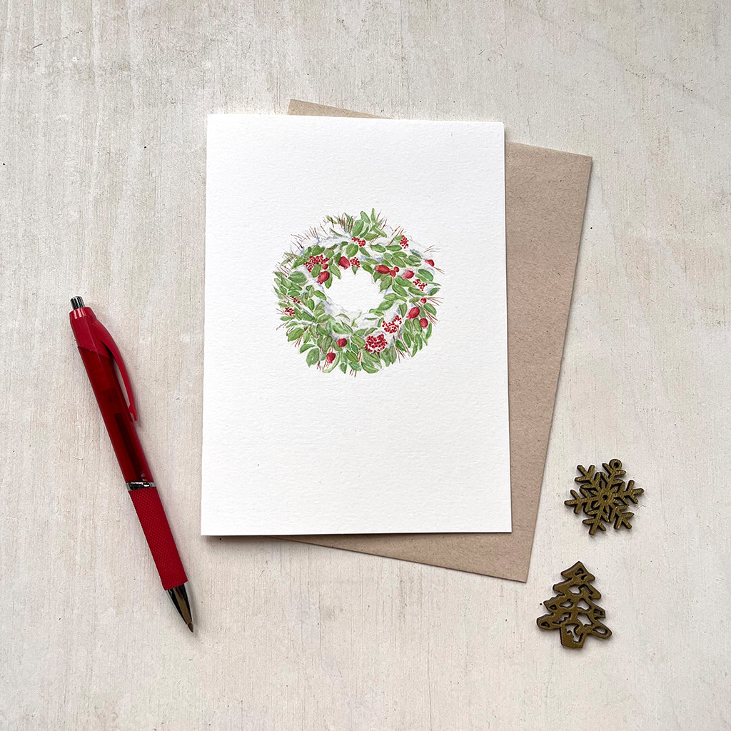 A Christmas card with a delicate watercolour painting of a snow-covered holiday wreath made of green leaves, red fruit and berries.