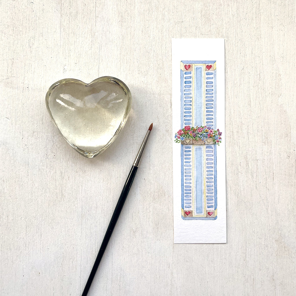 A paper bookmark featuring a delicate watercolour painting of a window box filled with colourful flowers. Behind the window box is a blue shutter adorned with hearts.