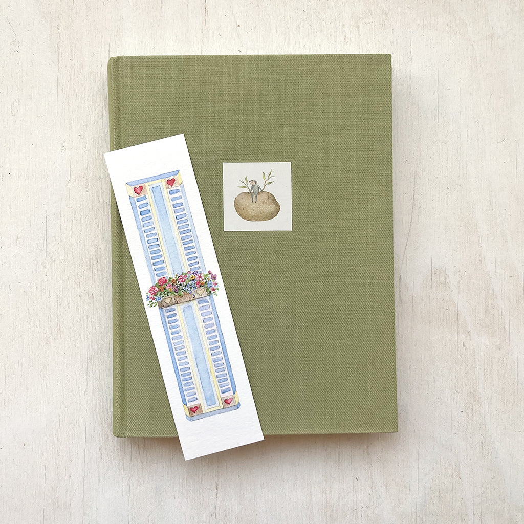 A paper bookmark featuring a delicate watercolour painting of a window box filled with colourful flowers. It is sitting on top of a book.