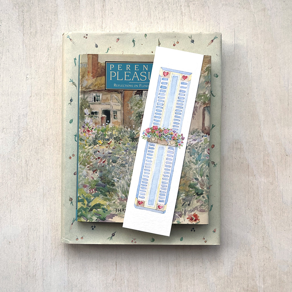 A paper bookmark featuring a delicate watercolour painting of a window box filled with colourful flowers sitting on top of a stack of books.